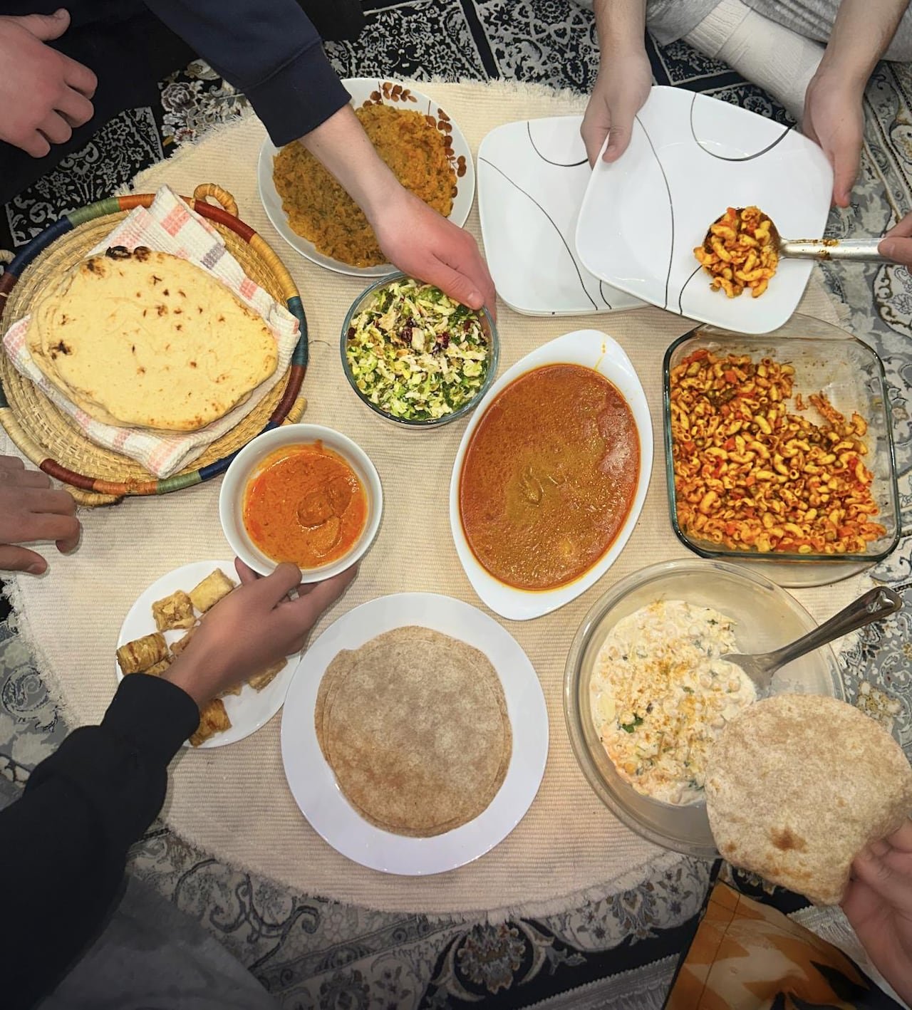 default-225 A bird’s eye view of several hands reaching for food, such as butter chicken, macaroni salad, roti and baklava.