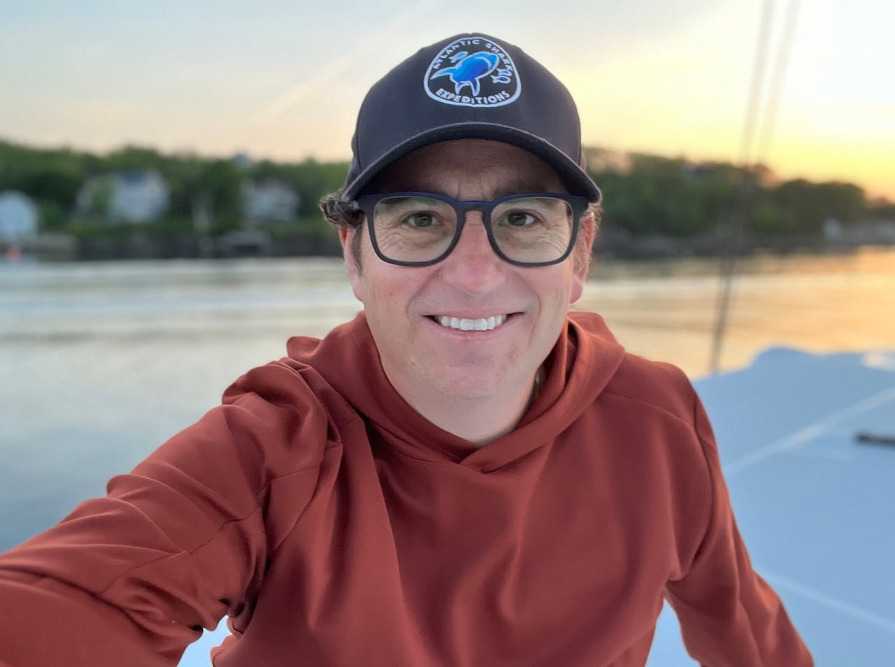 default-37 A smiling man with square glasses. He is wearing a baseball hat and an orange hoodie sitting on a boat.